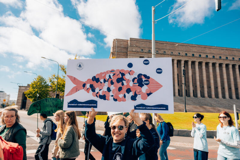 Ihmisiä kävelemässä eduskuntatalon edessä ja kantamassa kalakylttejä