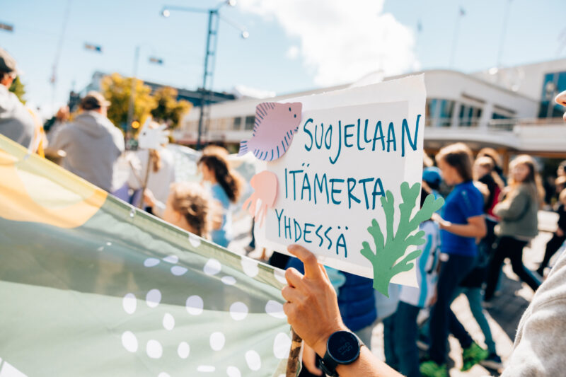 ihmisiä kävelemässä kulkueessa kyltin kanssa, jossa lukee suojellaan Itämerta yhdessä