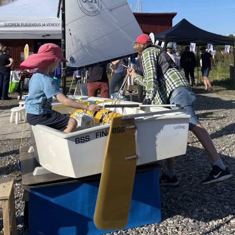Två personer i rosa kepsar provar en liten segeljolle uppställd på ställning, med segel och ett gult roder synligt, medan tält och folk syns i bakgrunden under ett utomhusevent i solsken.