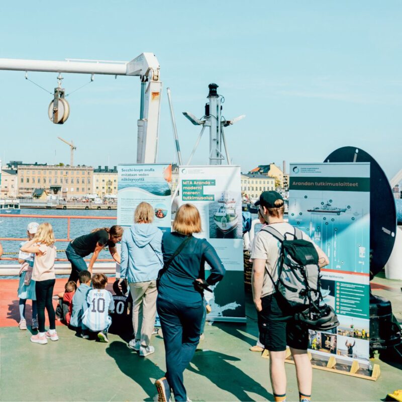 Forskningfartyget Arandas soldäck med en grupp besökare, främst barn och vuxna, som står och läser informationsskyltar och affischer om forskningen; hamn- och stadssiluett i bakgrunden under klarblå himmel.