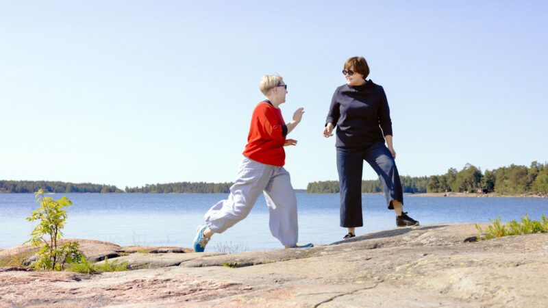 Kaksi ihmistä leikkii ja kävelee kallioilla merenrannalla aurinkoisena päivänä; vasemmalla punapaitainen henkilö hypähtää oikealla olevaa tummiin vaatteisiin pukeutunutta henkilöä kohti, taustalla tyyni Itämeri ja saaret.
