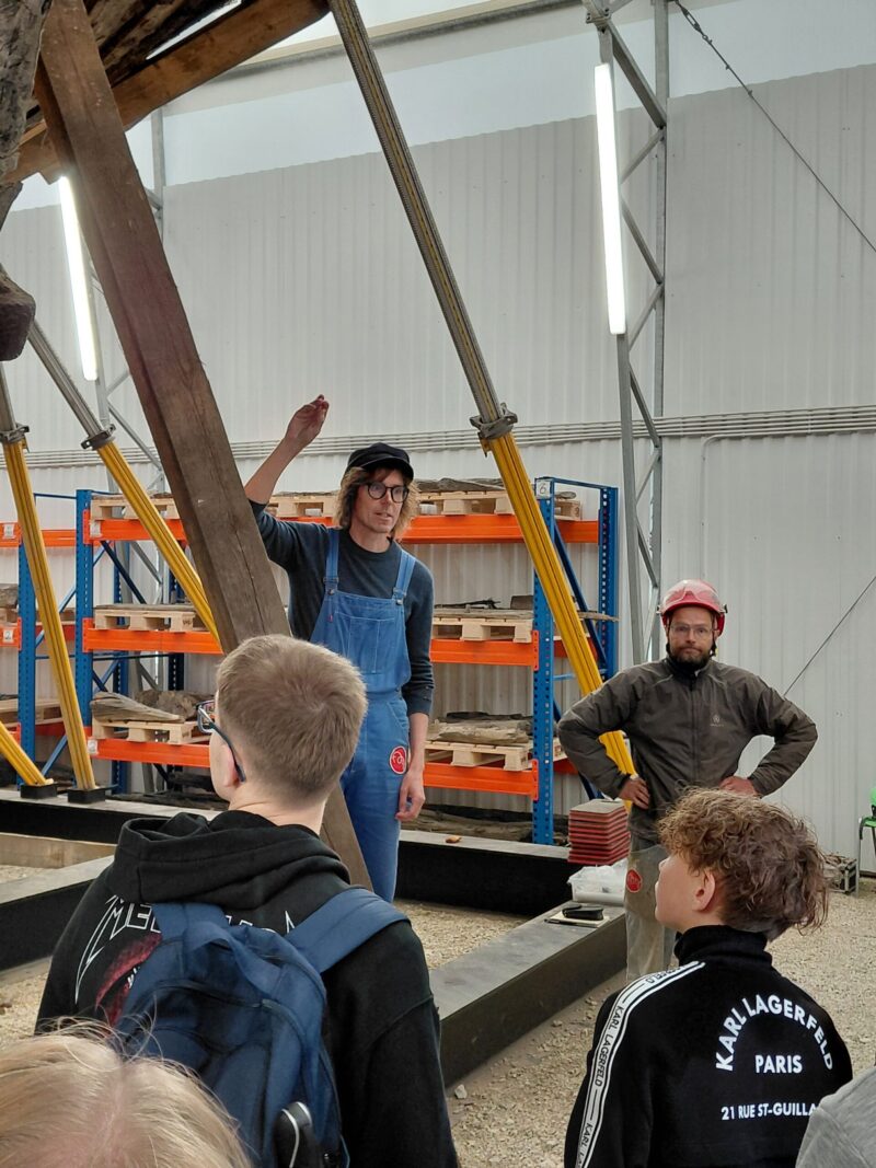 Restaureerimistöökojas kaks tööriietes meest – üks sinistes tööpükstes ja nokamütsiga käsi õhku tõstmas, teine punase kiivriga käed puusas – seletavad ees seisvatele noortele seljakottidega; taustal riiulid, tööriistad ja suured puittalad toestusvöödega.