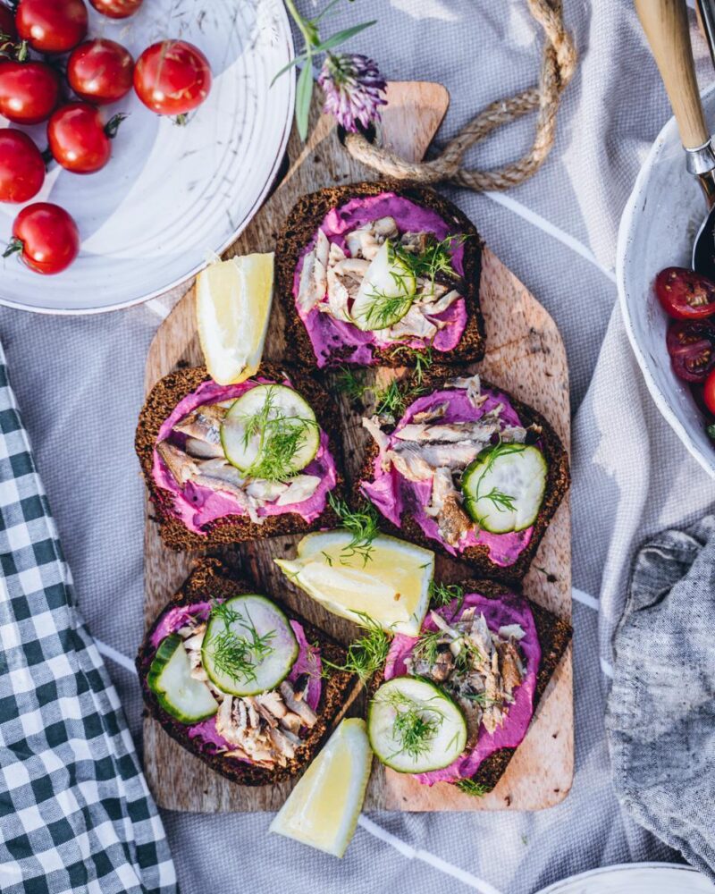 Savumuikkuleipiä, joissa on punajuuritahnaa, kurkkuviipaleita, tilliä ja sitruunaviipaleita puisella leikkuulaudalla, ympärillä kirsikkatomaatteja ja ruokailuvälineitä.