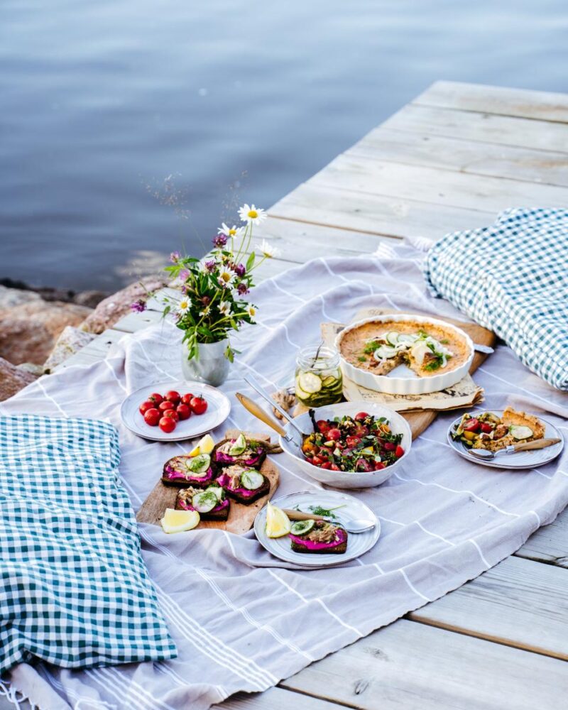 Piknik-tyylinen ateria kalliolla meren rannalla, jossa on erilaisia kaloista ja kasviksista valmistettuja ruokia, kukkakimppu ja sinivalkoruudulliset tyynyt.