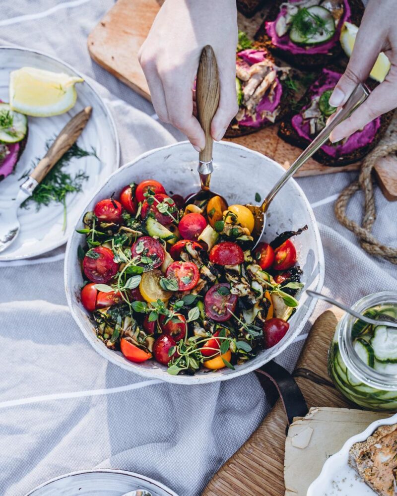Kädet tarjoilemassa värikästä salaattia, jossa on kirsikkatomaatteja, yrttejä ja siemeniä, pöydällä myös muita ruokia ja juomia.