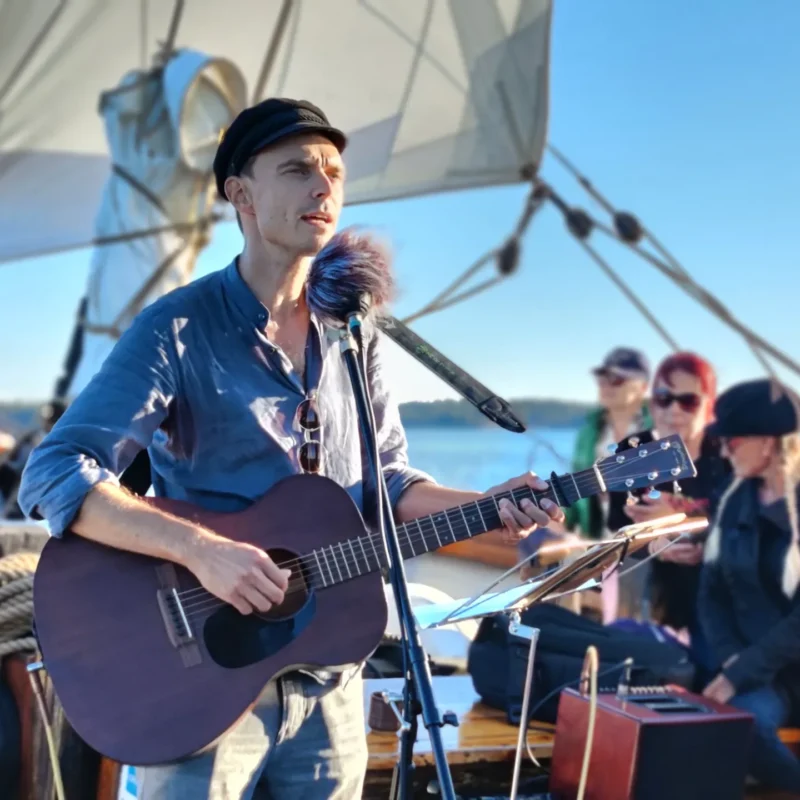 Mies sinisessä paidassa ja lakissa soittaa akustista kitaraa ja laulaa mikrofonin edessä purjeveneen kannella, taustalla istuu yleisöä ja näkyy sininen meri ja taivas.