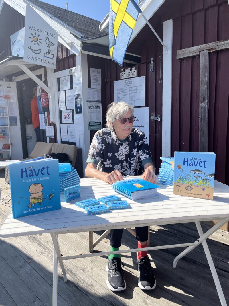 Lasse Åberg med solglasögon i blommig skjorta sitter bakom ett vitt bord på en träbrygga utanför en röd träbyggnad med hissad svensk flagga och visar upp flera exemplar av en barnbok med titeln "Havet".